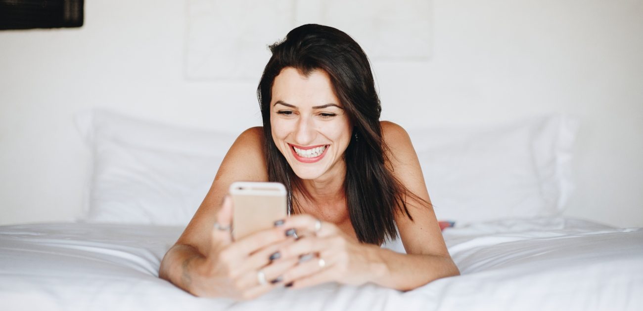 امرأة تنظر مبتسمة إلى هاتفها وهي مسلتقية على السرير