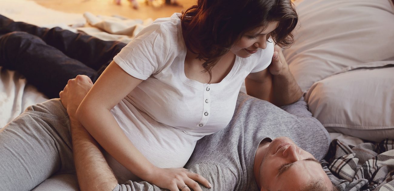 امرأة حامل تجلس إلى جانب زوجها المستلقي على السرير بسعادة