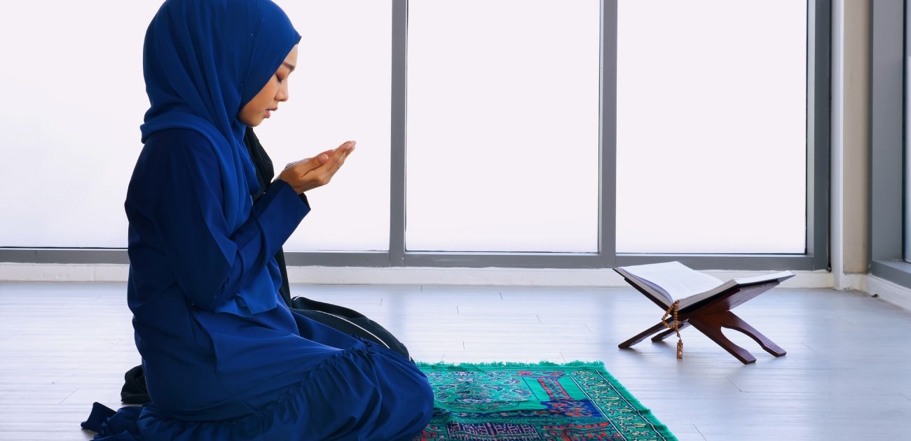امرأة مسلمة جالسة على سجادة الصلاة وترفع يديها للدعاء