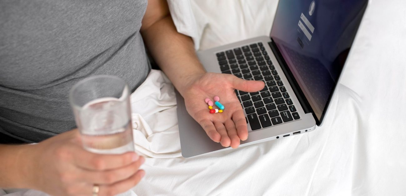امرأة تجلس في سريرها تمسك في يدها مجموعة من حبوب الأدوية وكوب من الماء في اليد الأخرى وأمامها حاسوب محمول