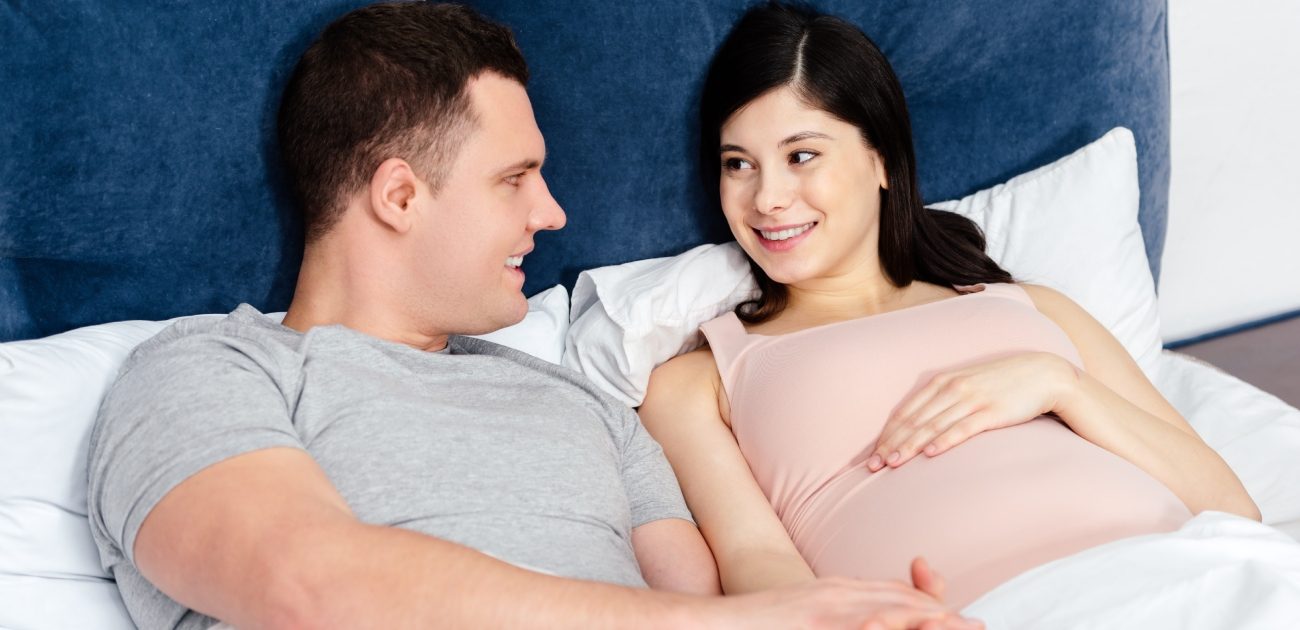 امرأة حامل بجانب زوجها على السرير يبتسمان وينظران إلى بعضهما بسعادة