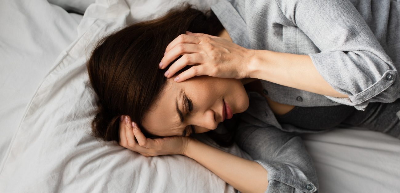امرأة مستلقية على السرير تغمض عينيها بشدّة وتضع يديها على رأسها
