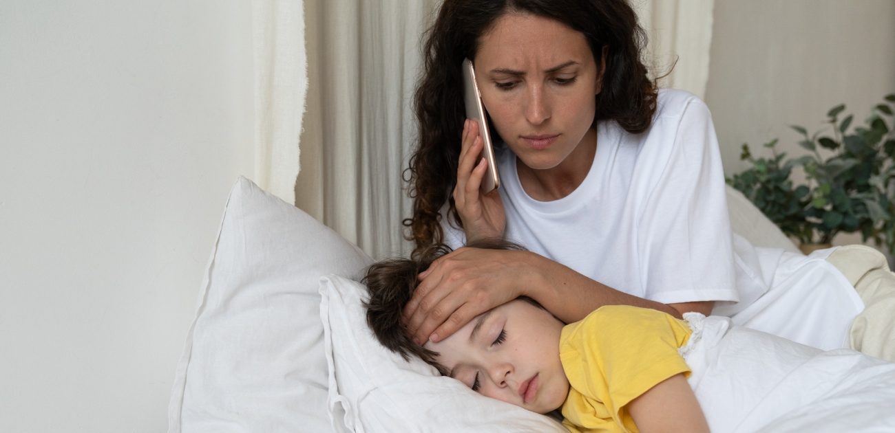 طفل نائم في السرير ووالدته تضع يدها على رأسه لتستشعر درجة حرارته وتتكلّم على هاتفها المحمول