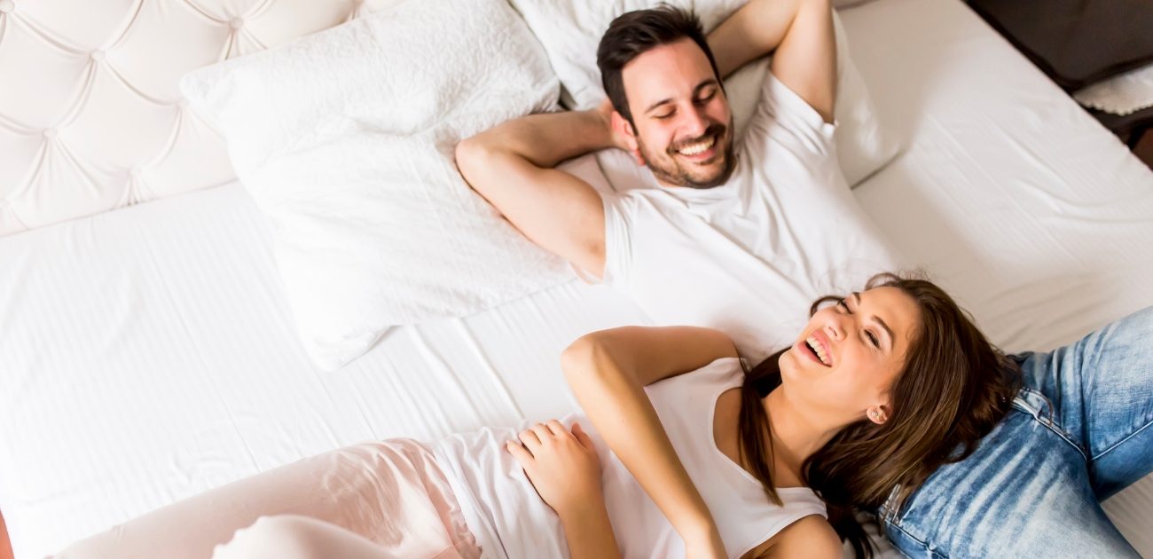 رجل مستلقٍ على السرير وزوجته مستلقية تضع رأسها على بطنه وهما يضحكان