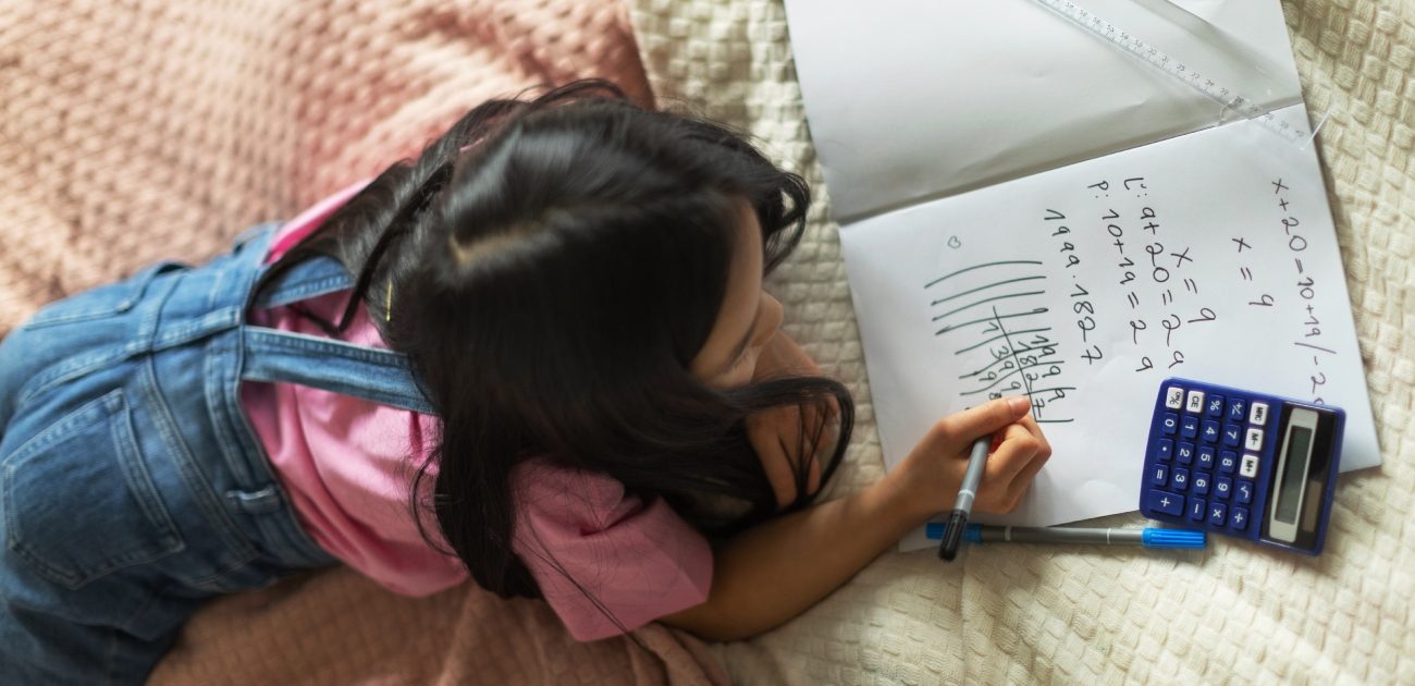 طفلة مستلقية على بطنها على السرير وتحلّ مسألة رياضيات على دفتر وبجانبها آلة حاسبة