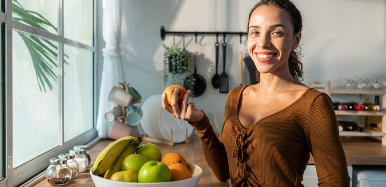 امرأة مبتسمة تمسك برتقالة وأمامها إناء يحتوي أنواع مختلفة من الفواكه