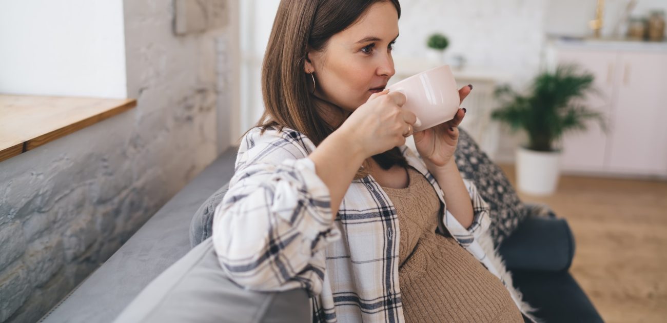 امرأة حامل تجلس على الكنبة وتشرب مشروبًا ساخنًا