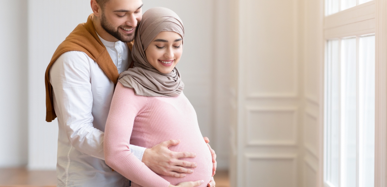 زوج يُعانق زوجته الحامل من الخلف ويضع يديه على بطنها