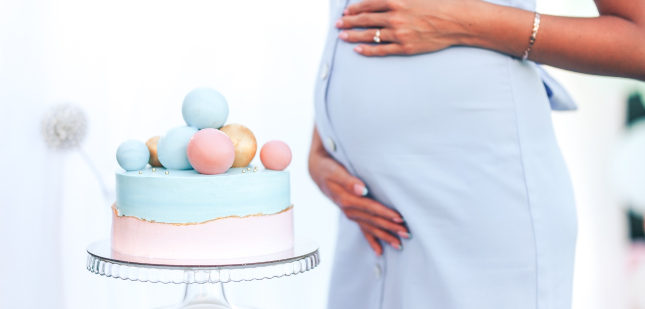 امرأة حامل تضع يديها على بطنها وبجانبها قالب حلوى مزيّن باللونين الأزرق والزهري
